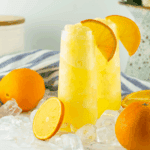 Glass of frozen mimosa slush made with orange juice and champagne, topped with an orange slice and served with a straw.