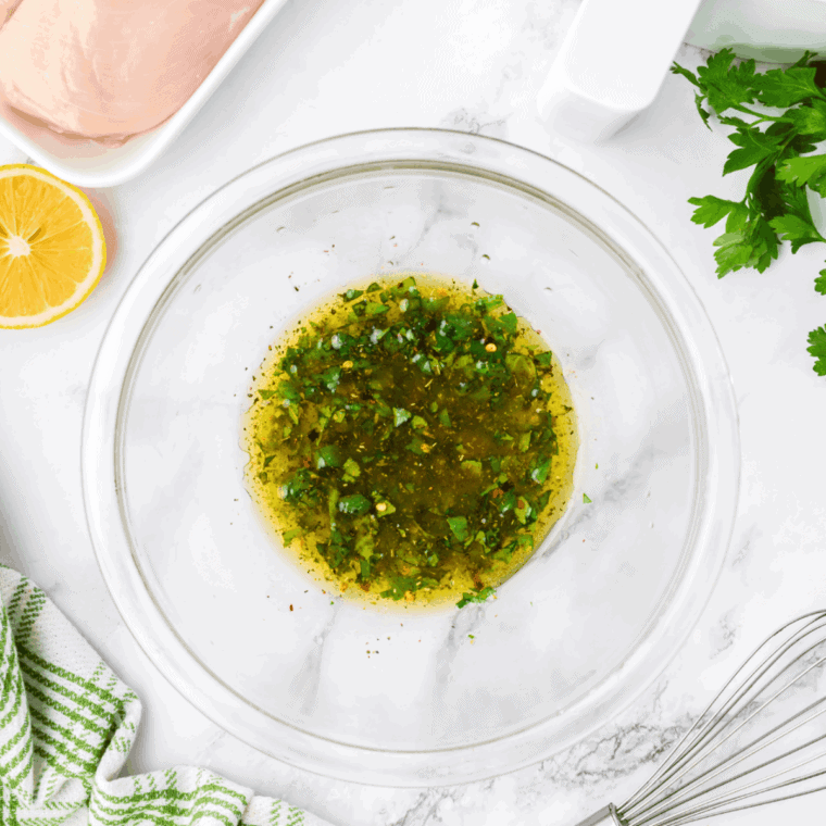 Raw chicken breasts marinating in a clear, oil-based Italian Marinade, flecked with green herbs and spices, in a zipper bag or glass dish.