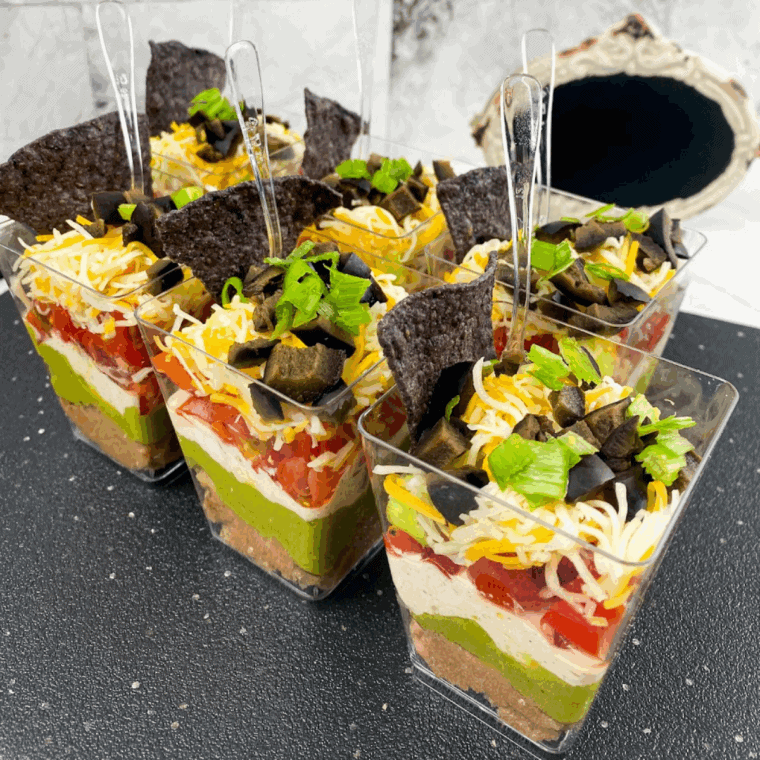 Individual 7 Layer Dip Cups with beans, guacamole, seasoned sour cream, shredded cheese, tomatoes, olives, and green onions.