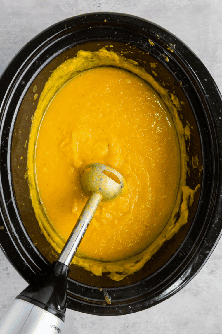 Immersion blender pureeing butternut squash soup in the slow cooker until creamy and smooth.