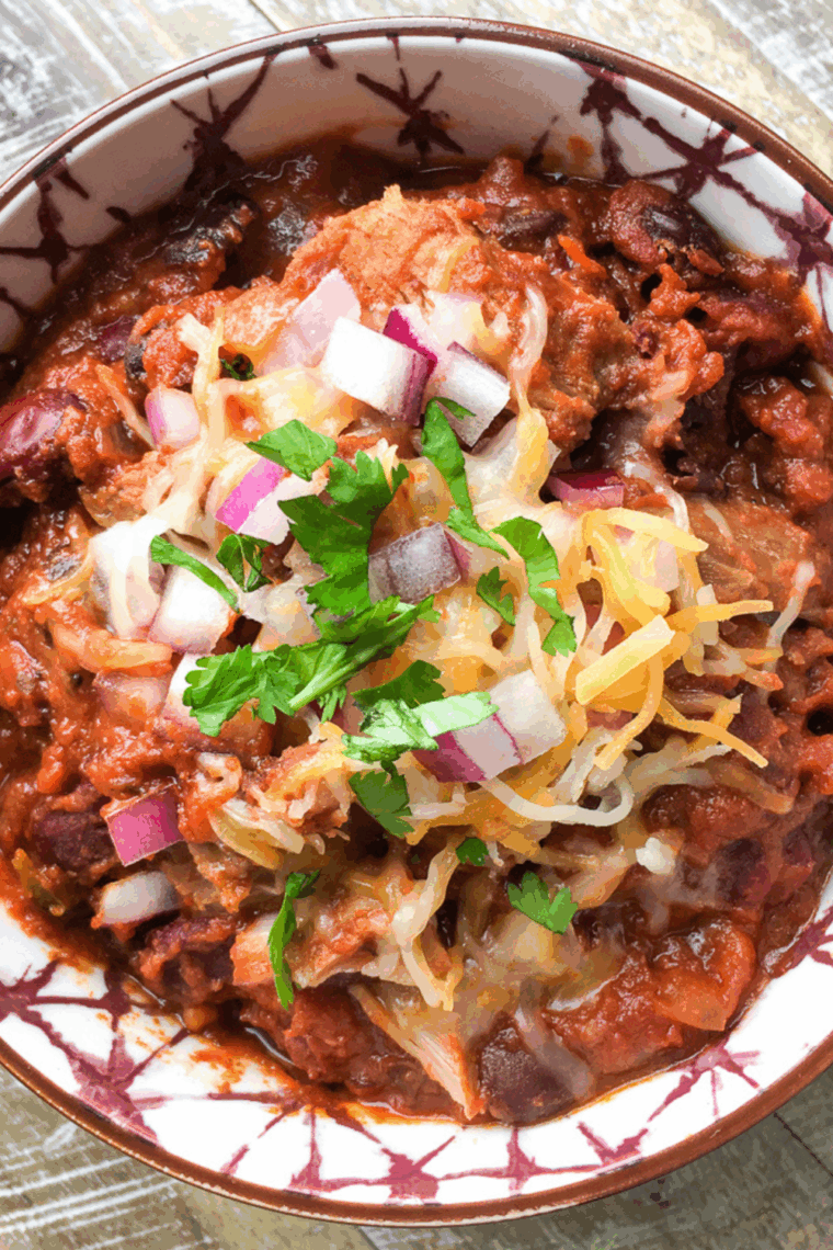 Slow Cooker Pulled Pork Chili with shredded pork, beans, diced tomatoes, and spices in a bowl, topped with cheese and fresh cilantro.