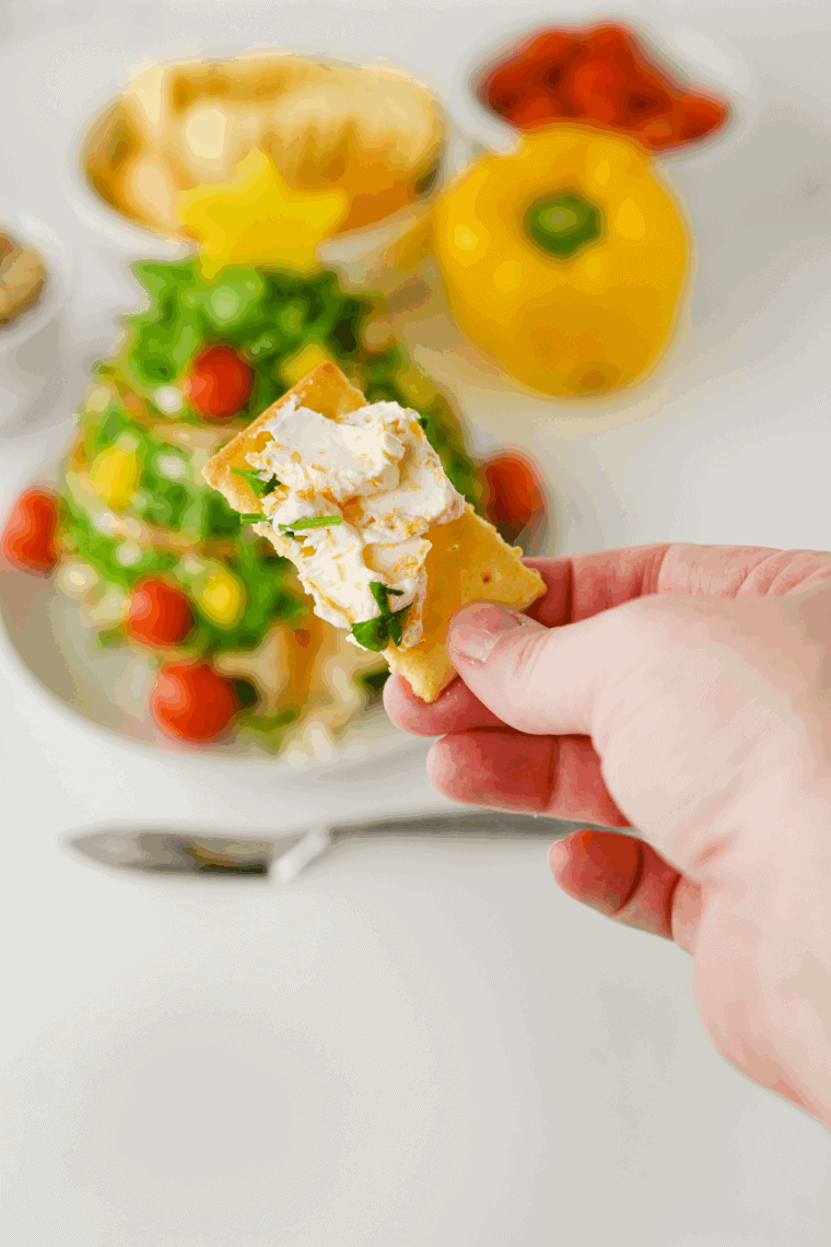 A festive Christmas Tree Cheese Ball coated in chopped herbs, decorated with red bell pepper 'ornaments,' and served on a platter with assorted crackers.