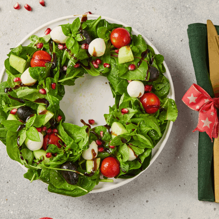 A festive Christmas Wreath Salad made with a ring of mixed green lettuce, decorated with red cherry tomato halves, white mozzarella balls, and yellow star-shaped vegetables.