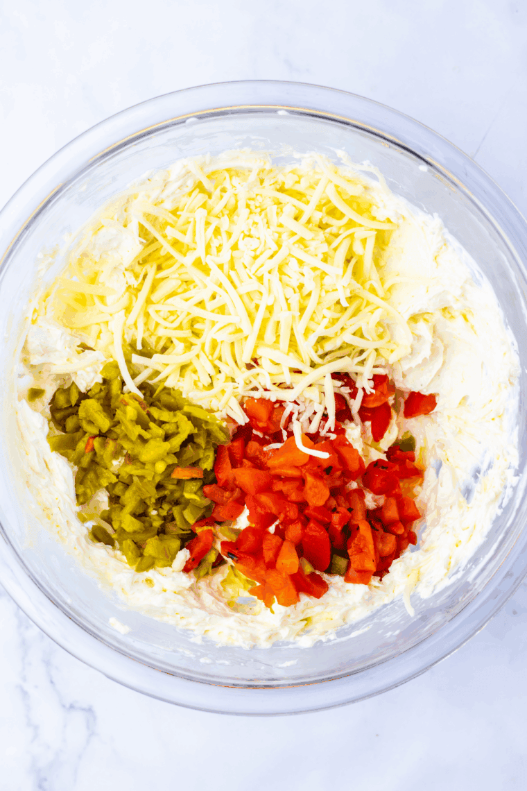 Folding diced red peppers, chopped jalapeños, and shredded cheese into creamy mixture in a bowl.