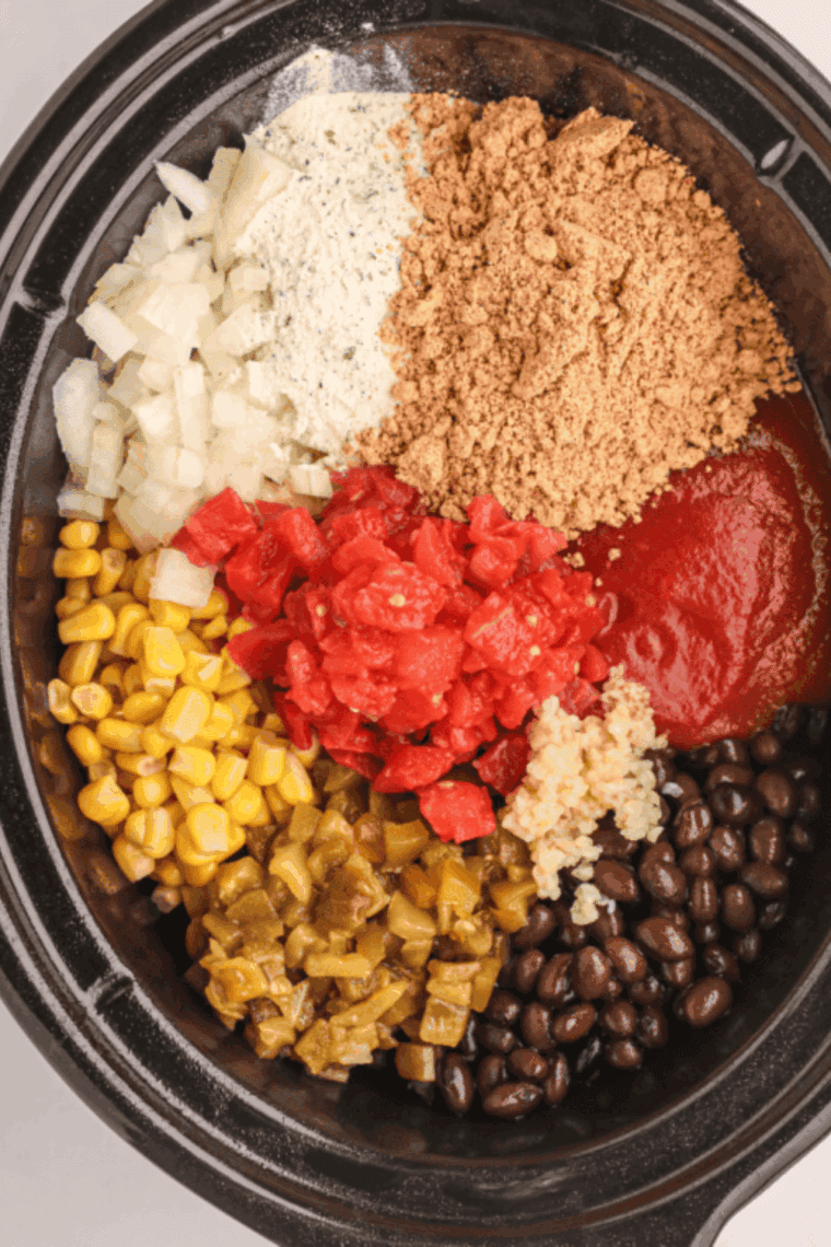 Ingredients layered in a slow cooker with raw chicken breasts, beans, corn, chiles, onions, garlic, and spices.