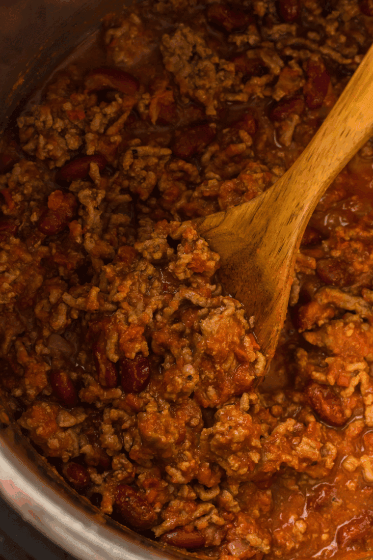 A steaming bowl of thick, beefy George’s Chili topped with a generous sprinkle of shredded cheddar cheese and finely diced red onions, served with a side of saltine crackers.
