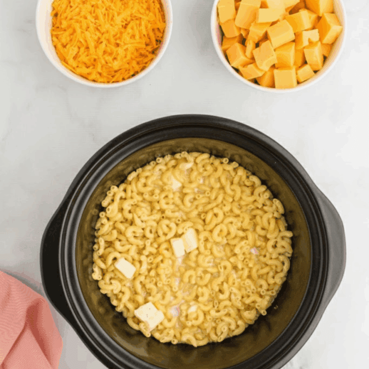 Stirring shredded sharp cheddar and cubed American cheese into the pasta mixture in a black Crockpot.