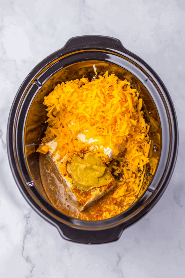 Shredded cheddar cheese evenly layered over cream cheese in the slow cooker.