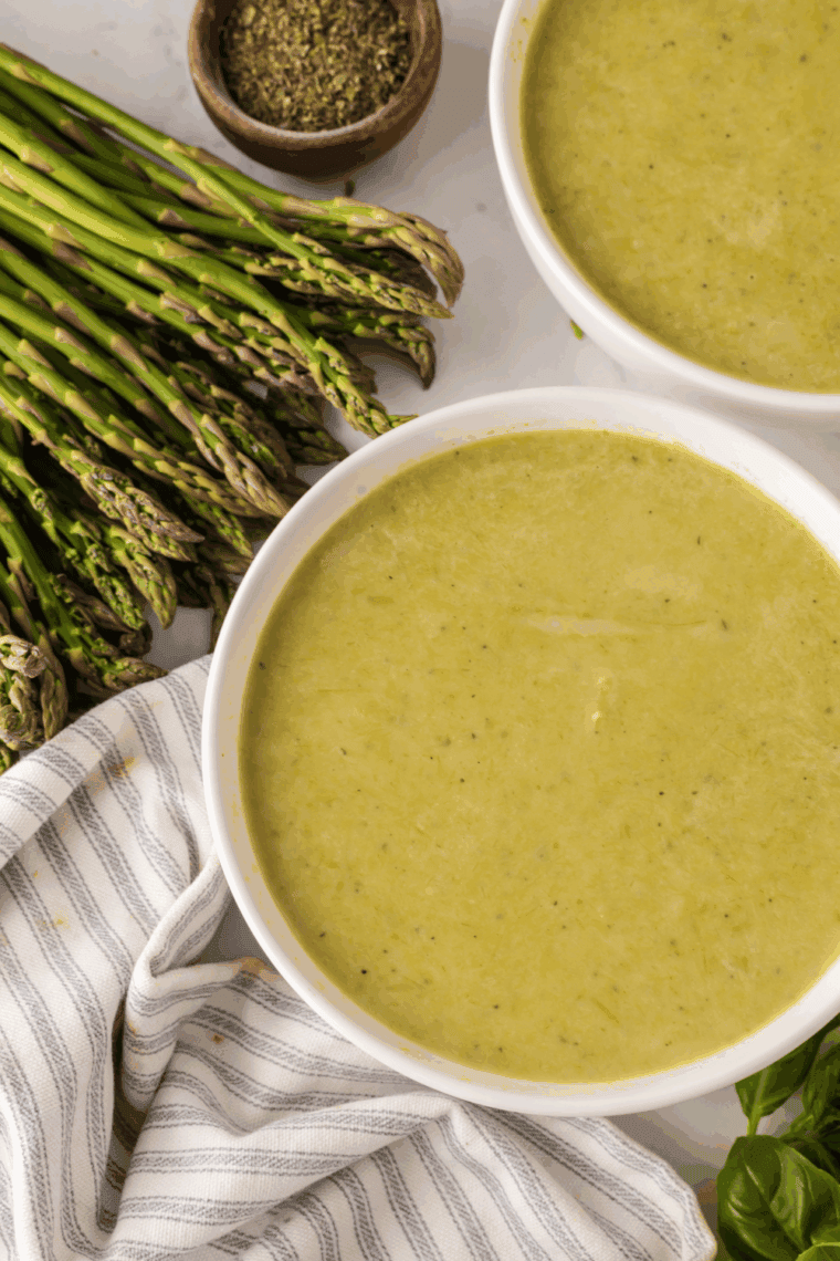 A white ceramic bowl filled with vibrant green, creamy asparagus soup, garnished with a swirl of fresh cream and a few tender roasted asparagus tips.
