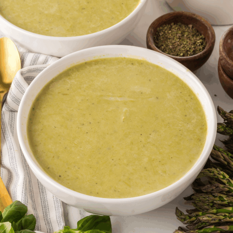 A steaming bowl of vibrant green asparagus soup garnished with fresh cream swirls and cracked black pepper.