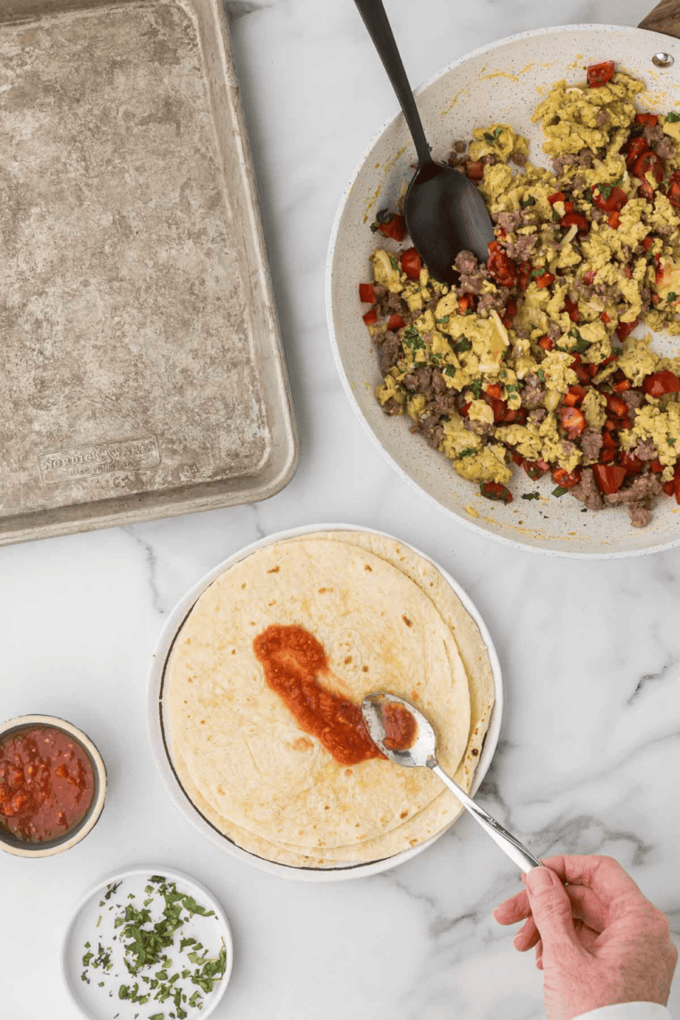 Placing scrambled egg mixture onto tortillas, spreading salsa, and rolling into burritos.
