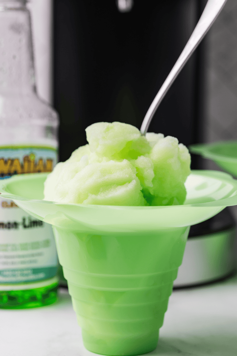 Pale yellow-green lemon-lime Italian ice in a clear glass, icy texture, garnished with a lime wedge on a white countertop.