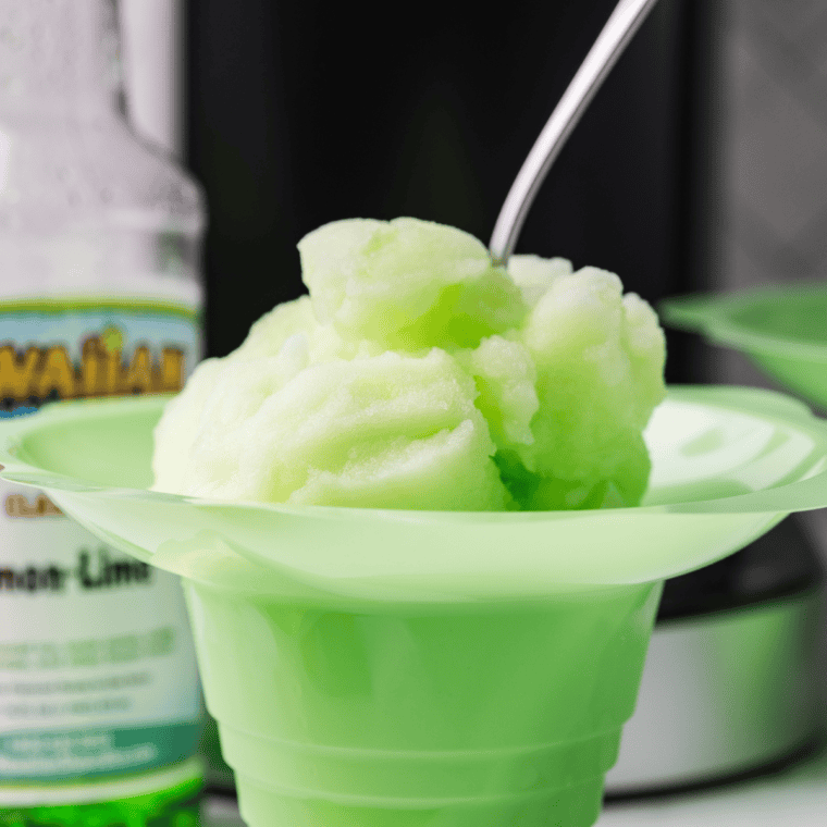 A glass of homemade lemon-lime Italian ice, pale yellow-green with icy texture, garnished with a lime wedge, on a white countertop.