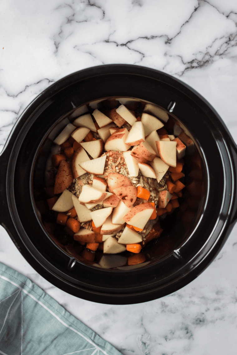 Chopped onions, carrots, potatoes, and celery arranged in slow cooker with herbs.