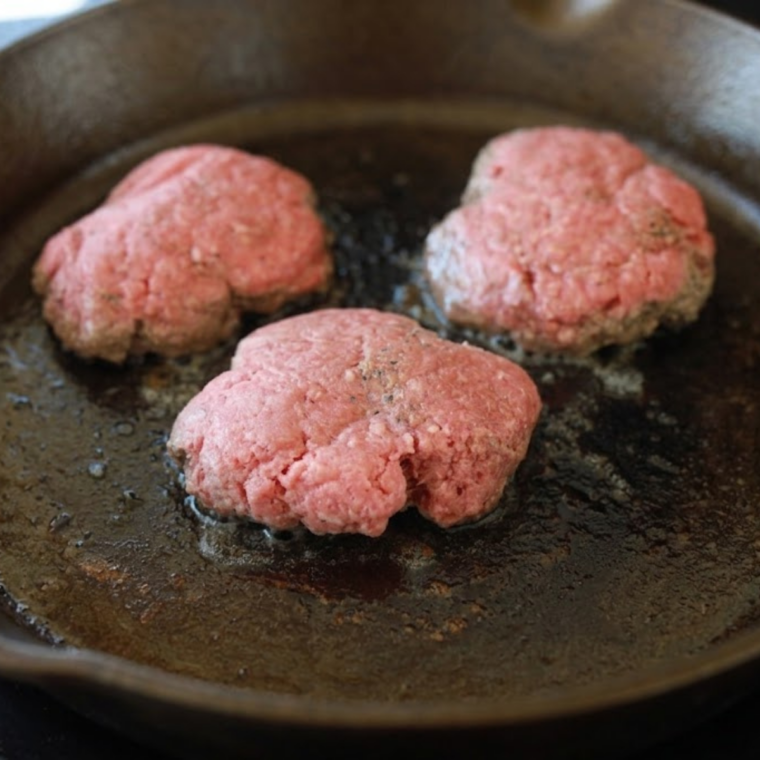 Smash burger being pressed flat in sizzling cast iron skillet creating crispy edges.