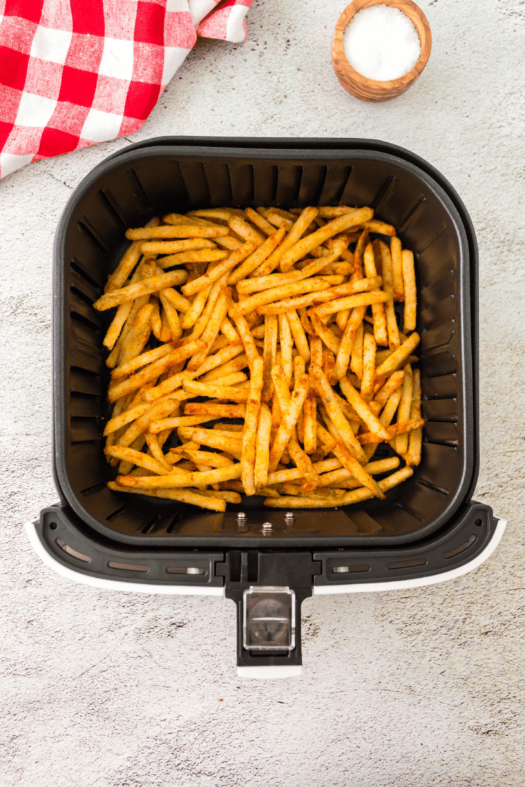 Homemade French fry seasoning being poured into an airtight spice jar, ready for storage, with crispy hot fries in the background being sprinkled with seasoning for a savory flavor boost.
