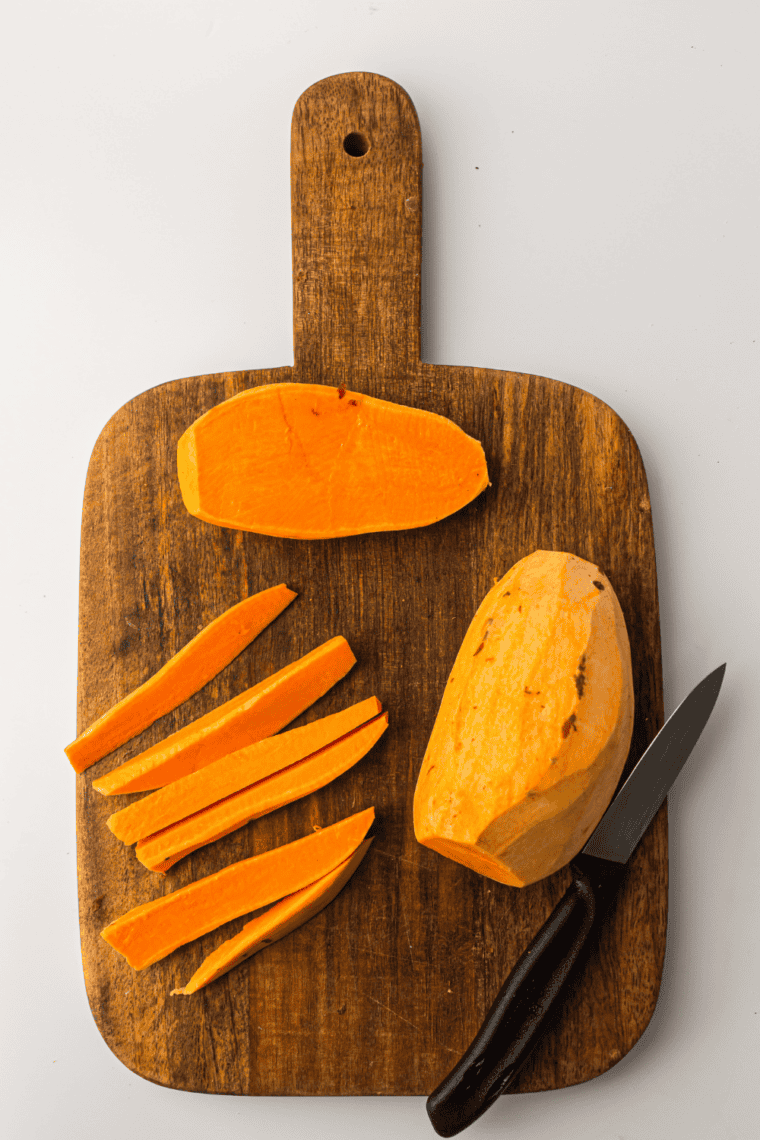 Sweet potatoes being peeled and cut into uniform fry-sized sticks on a cutting board.