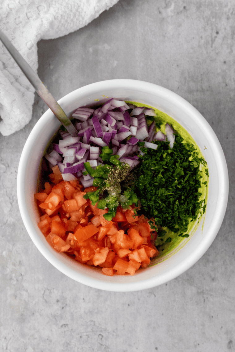 Folding diced red onion, jalape&ntilde;o, cilantro, and Roma tomatoes into mashed avocado for copycat Chipotle guacamole