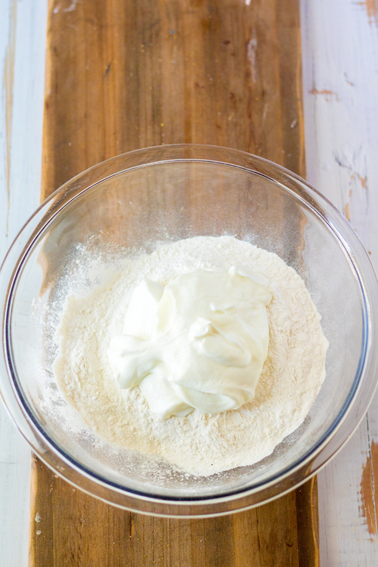 Mixing self-rising flour and Greek yogurt in a bowl with a wooden spoon until a shaggy dough forms.
