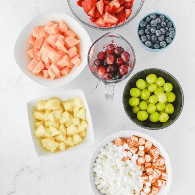 Ingredients for a rainbow fruit tray laid out in bowls: strawberries, cantaloupe, pineapple, grapes, blueberries, marshmallows, and Rolos.