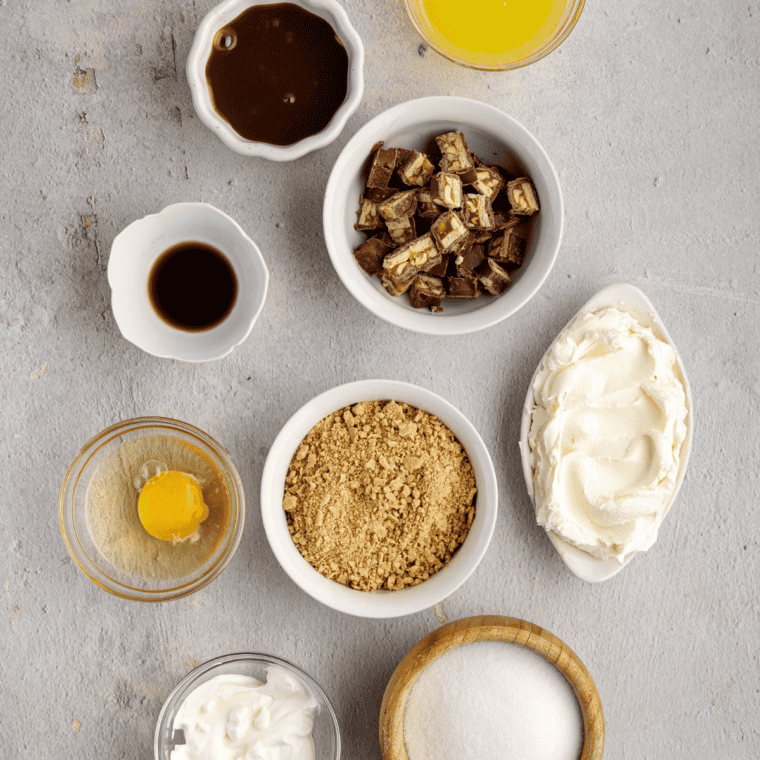 Ingredients for air fryer Snickers cheesecake: cream cheese, Snickers bars, graham crackers, eggs, sour cream, caramel, and peanuts