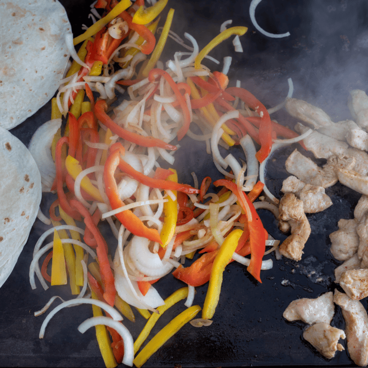 Bell peppers and onions charring beside cooked chicken on a Blackstone griddle