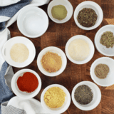 Granulated garlic, dried oregano, basil, lemon peel, and red pepper flakes measured in bowls for garlic herb seasoning