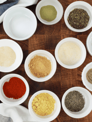 Granulated garlic, dried oregano, basil, lemon peel, and red pepper flakes measured in bowls for garlic herb seasoning