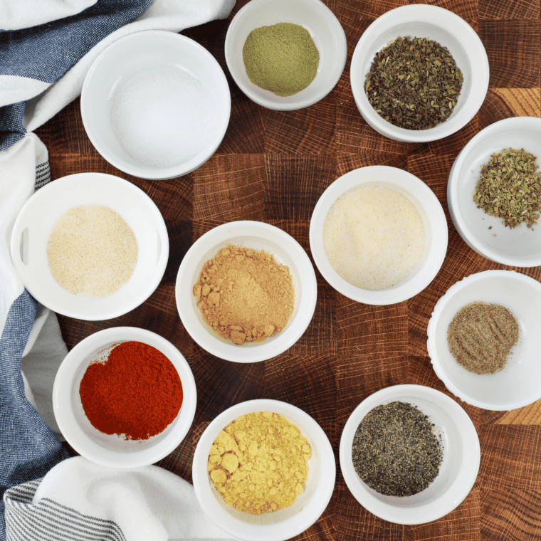 Granulated garlic, dried oregano, basil, lemon peel, and red pepper flakes measured in bowls for garlic herb seasoning.