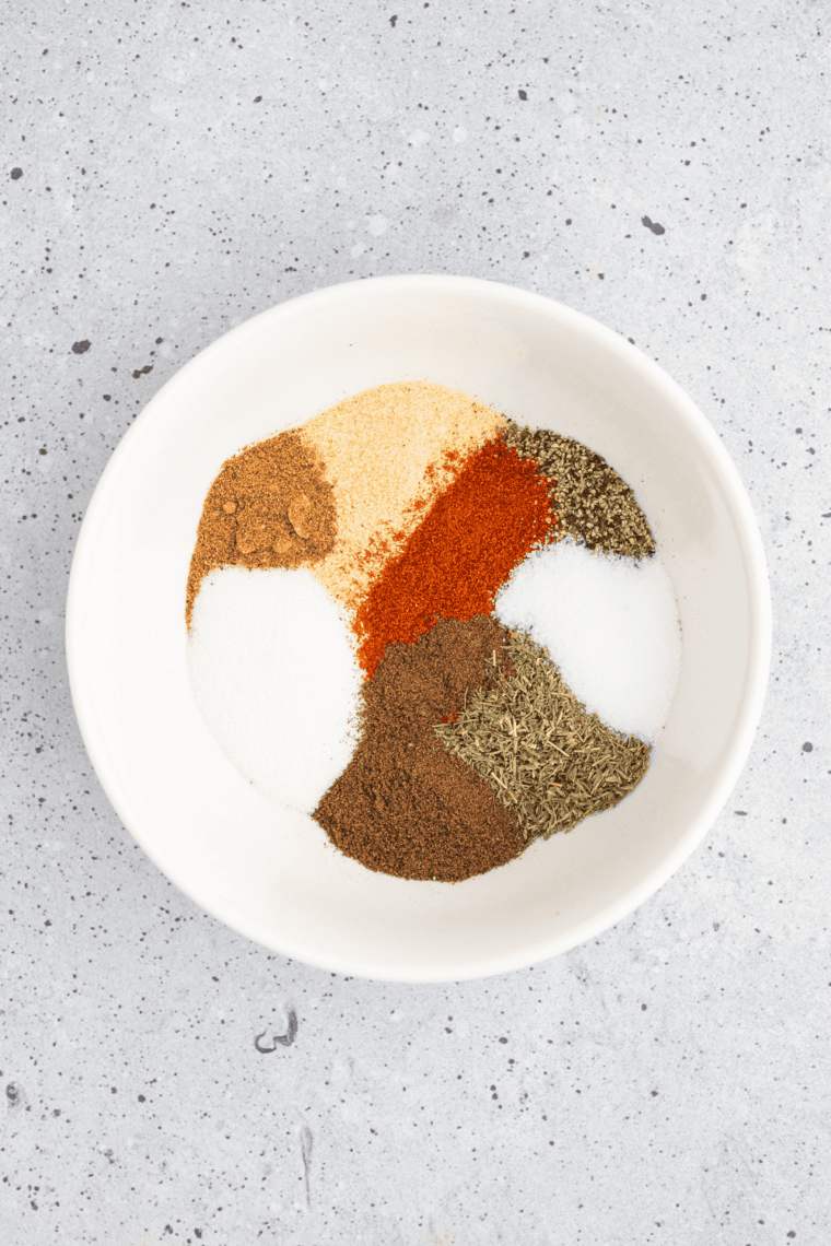 Granulated garlic, dried oregano, basil, lemon peel, and red pepper flakes measured in bowls for garlic herb seasoning