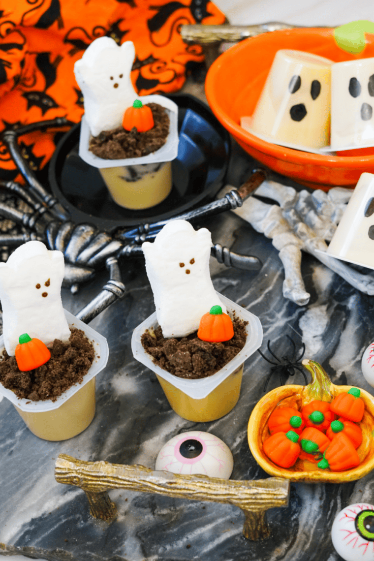 Three Halloween pudding cups with marshmallow ghosts and candy pumpkins on a dark table