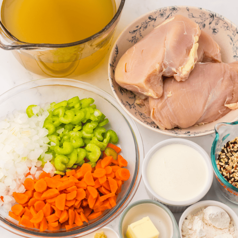 Ingredients for slow cooker chicken and wild rice soup arranged on a counter, including chicken, wild rice, carrots, celery, onion, and broth