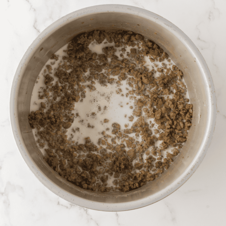Warm milk being poured into the sausage and flour roux while stirring in a skillet