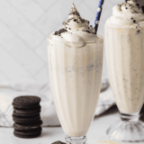 Copycat Chick-fil-A cookies and cream shake in a tall glass topped with whipped cream, crushed Oreo pieces, and a maraschino cherry