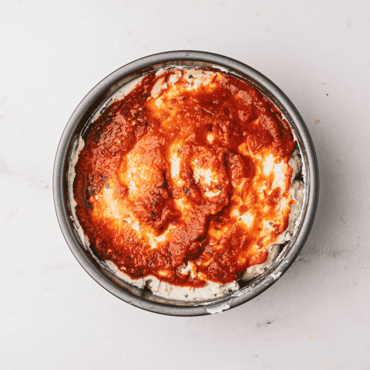 Pizza sauce being spread onto raw pizza dough with the back of a spoon