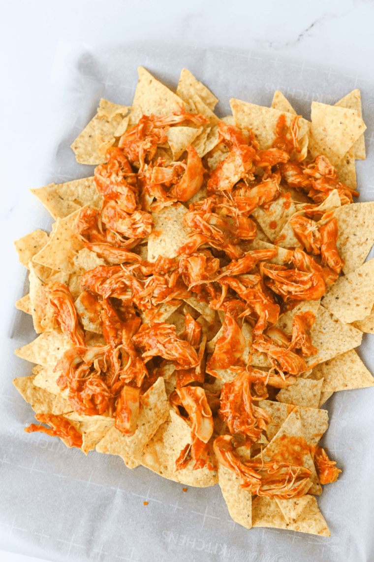 Tortilla chips on a sheet pan topped with shredded salsa chicken, corn, black beans, and shredded cheddar before broiling