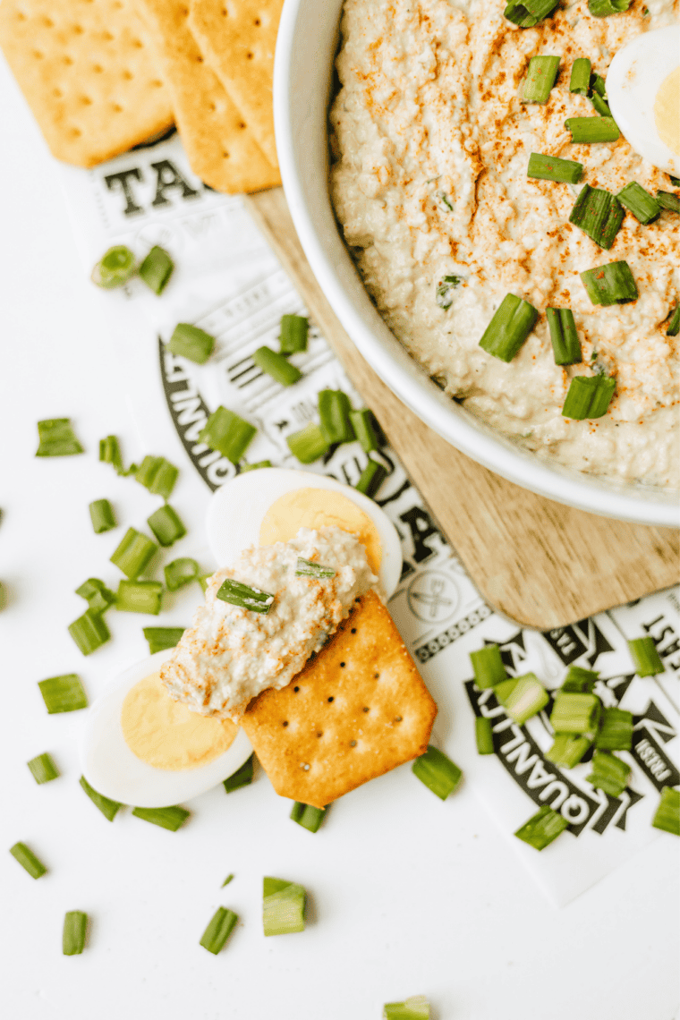 Deviled egg dip topped with paprika and green onions in a white serving bowl with Ritz crackers