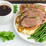 Sliced slow cooker tri tip on a wooden board with brown gravy and fresh rosemary