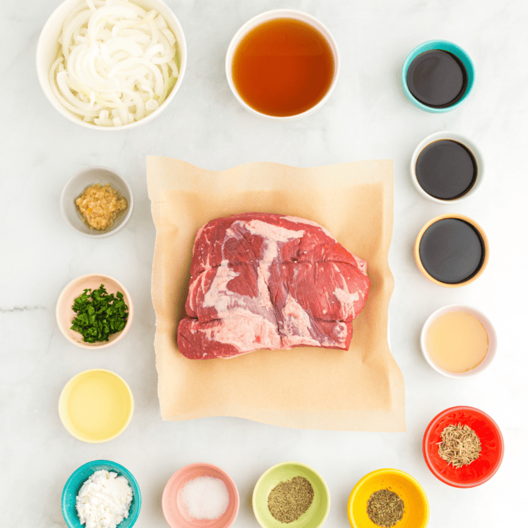 Tri tip roast, onion, garlic, beef broth, and spices arranged on a marble counter for slow cooker tri tip