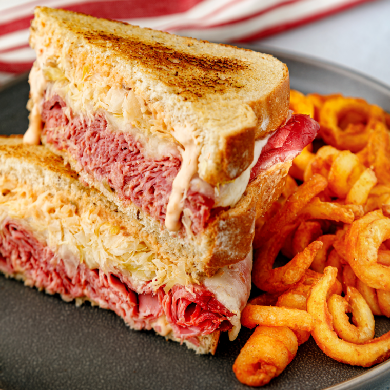 Several Reuben sandwiches grilling on a Blackstone griddle with corned beef warming beside them