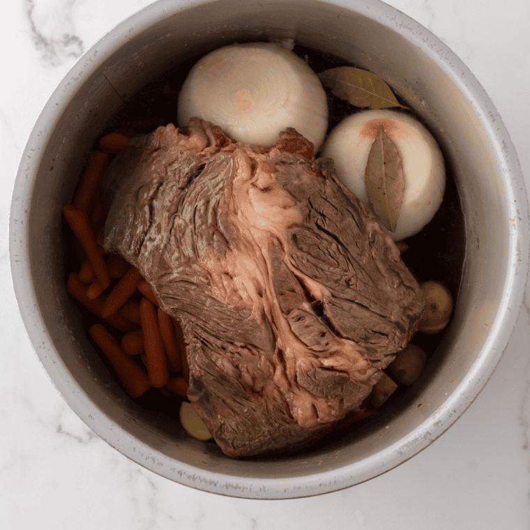 Seared chuck roast in a slow cooker surrounded by quartered onion, smashed garlic, bay leaves and beef broth